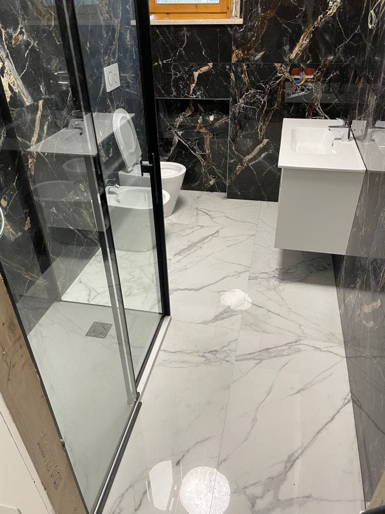 Bagno ristrutturato con rivestimento effetto marmo nero, pavimento effetto marmo bianco e box doccia in vetro